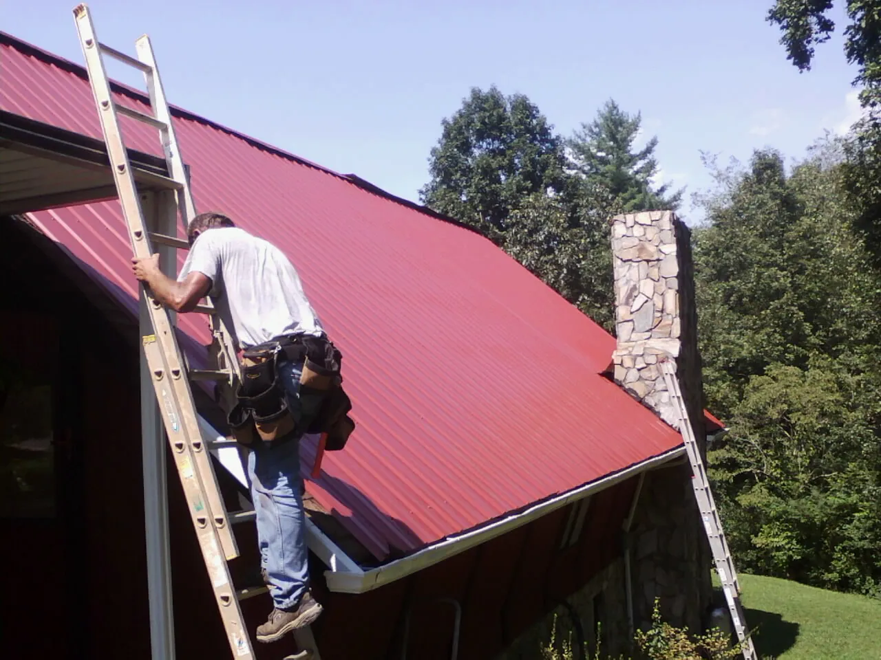 Roofer on a ladder beside a red metal roof and stone chimney — Soffit & Fascia Repair by Roofing Brothers in East Lyme